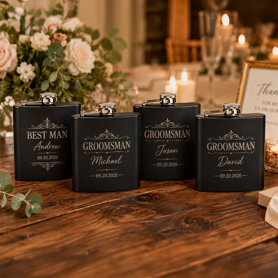 Set of four engraved black matte stainless steel flasks on a rustic wood table, including one Best Man flask for Andrew and three Groomsman flasks for Michael, Jason, and David, styled in a warm wedding setting with candles and floral décor. -- lxi-context:wedding