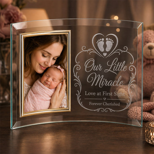 A curved glass photo frame displayed on a wooden table, featuring a color photo of a mother holding her sleeping newborn on the left in a gold-trimmed inset frame, and frosted engraved artwork on the right with baby footprints inside a heart, decorative flourishes, and the message “Our Little Miracle,” “Love at First Sight,” and “Forever Cherished,” set in a warm nursery-style background with soft lighting and a teddy bear. -- lxi-context:mothers-day