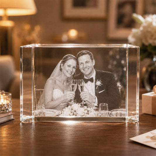 Wedding crystal photo keepsake block featuring an engraved bride and groom portrait raising champagne glasses, displayed on a wooden table in a warm cozy home setting with soft candlelight, floral accents, and gift box details -- lxi-context:wedding