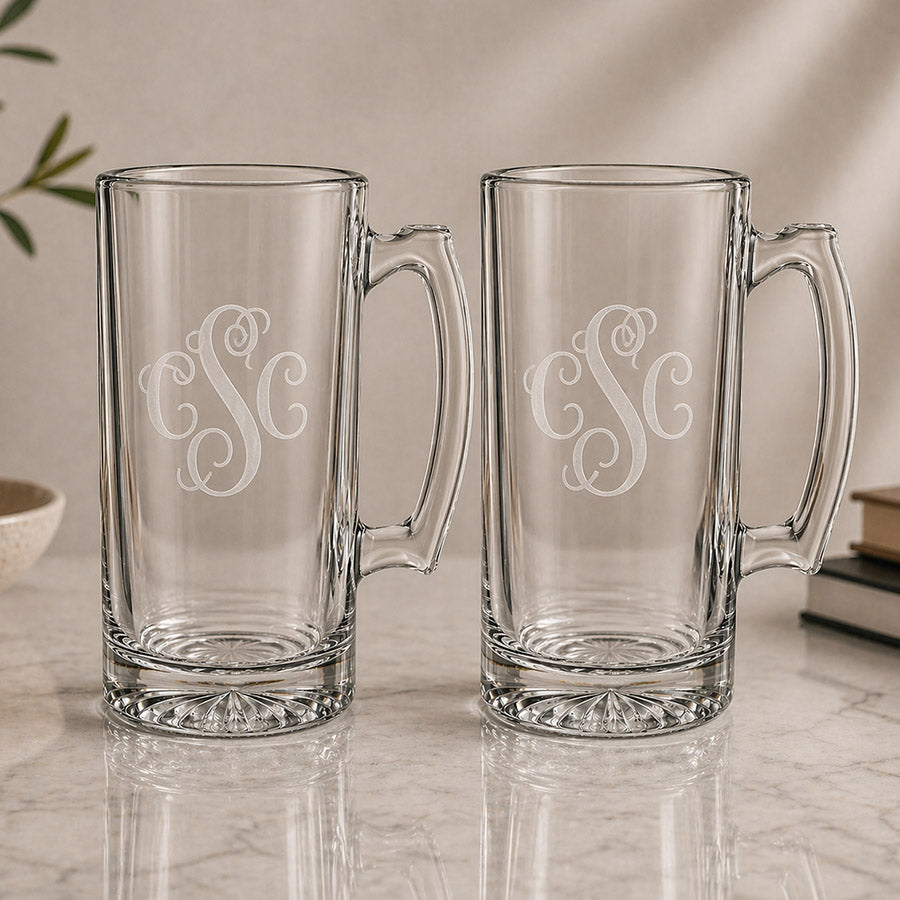 Set of two personalized clear glass beer mugs with matching engraved three letter monograms on a neutral tabletop display