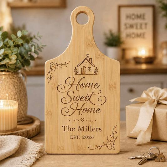 Engraved bamboo paddle cutting board displaying “Home Sweet Home” with a house icon, floral accents, and “The Millers EST. 2026,” styled on a countertop with a candle, wrapped gift, and house keys in a cozy home setting. -- lxi-context:house-warming