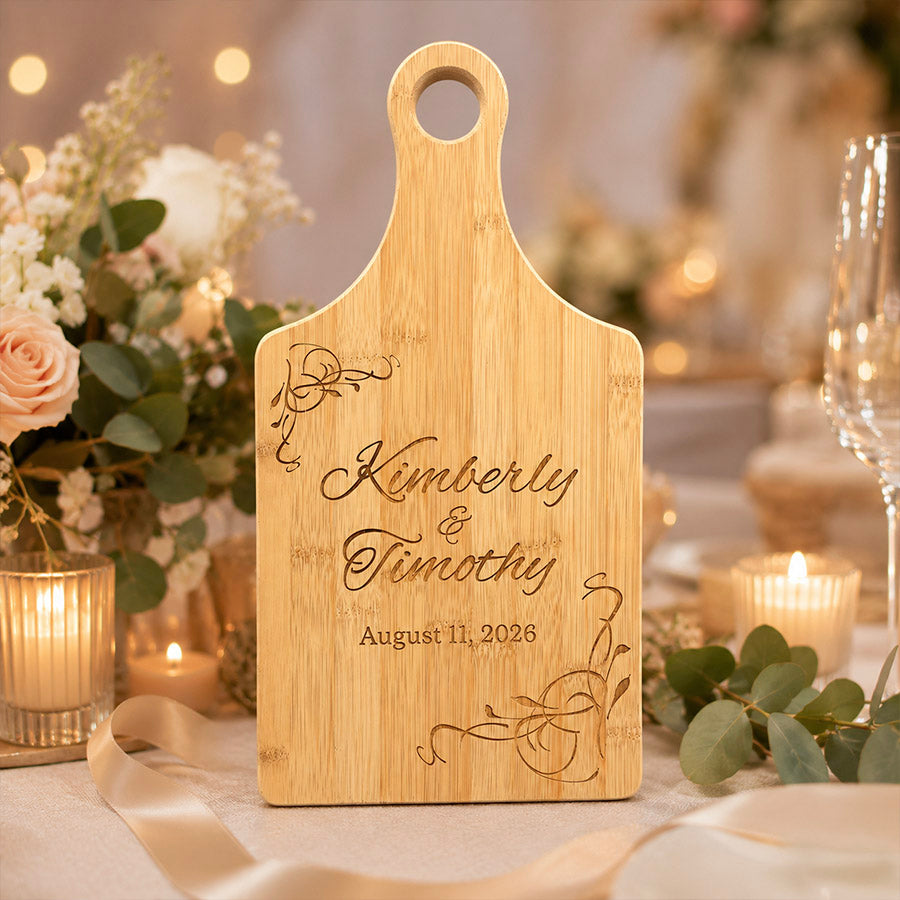 Engraved bamboo paddle cutting board featuring the names “Kimberly & Timothy” and the date “August 11, 2026,” displayed upright in a softly lit wedding setting with candles, blush flowers, ribbon, and eucalyptus accents. -- lxi-context:wedding
