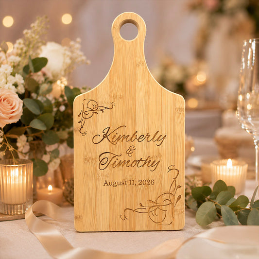 Engraved bamboo paddle cutting board featuring the names “Kimberly & Timothy” and the date “August 11, 2026,” displayed upright in a softly lit wedding setting with candles, blush flowers, ribbon, and eucalyptus accents. -- lxi-context:wedding