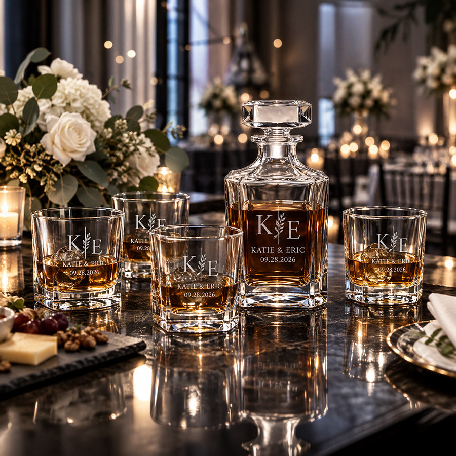 Personalized whiskey decanter set with four engraved glasses featuring K E monogram, Katie & Eric, and 09.28.2026 displayed on a reflective table in an elegant wedding reception setting -- lxi-context:wedding