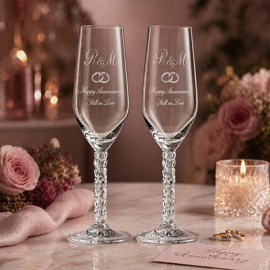 A pair of engraved champagne flutes with crystal-cut stems, each etched with the initials “R&M,” interlocking rings, and the message “Happy Anniversary Still in Love,” displayed on a marble surface in a romantic anniversary setting with pink roses, candlelight, wedding bands, and a handwritten anniversary card. -- lxi-context:anniversary