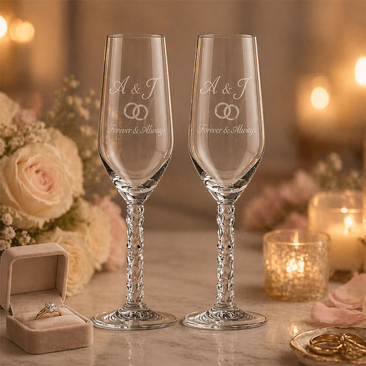 Two engraved champagne flutes with crystal-cut stems, each featuring the initials “A & J” above interlocking rings and the message “Forever & Always,” displayed in a romantic wedding setting with candles, blush flowers, a ring box, and wedding bands. -- lxi-context:wedding