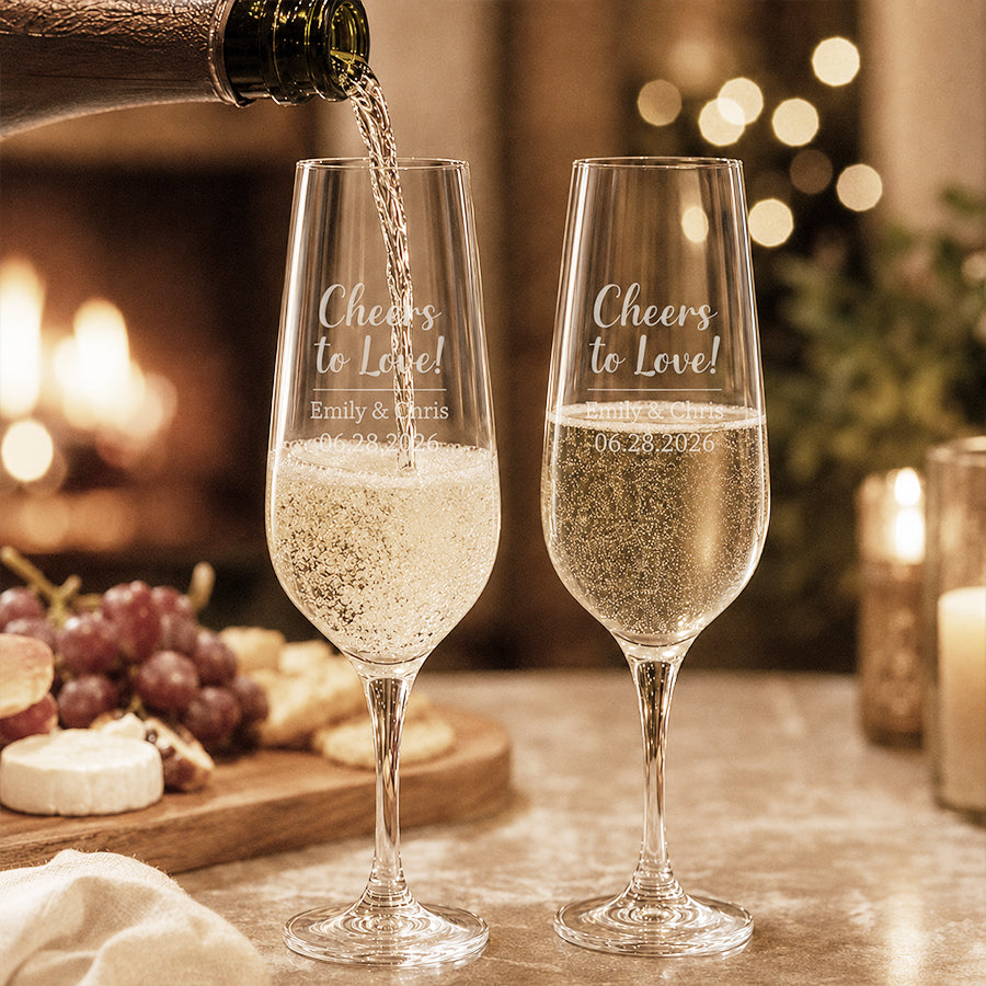 Personalized wedding champagne flutes engraved with "Cheers to Love!", couple names, and wedding date, with sparkling wine being poured beside a cozy fireplace and candlelit background